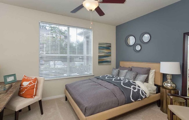Apartments for Rent in The Woodlands TX - Whispering Pines Ranch - Furnished Bedroom With Comfortable Bed, a Ceiling Fan, and a Desk With a Chair on Top of Carpet Flooring