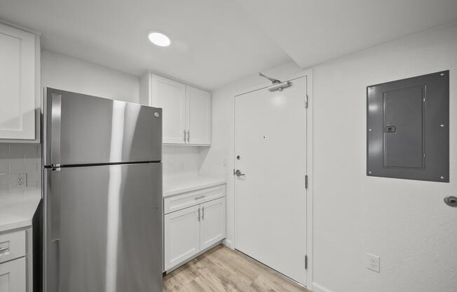 A modern kitchen with white cabinetry, a stainless steel refrigerator, and a solid door leading outside. The space features light-colored walls, a beige wood floor, and an electrical panel mounted on the wall near the entrance. Bright lighting enhances the clean and minimalist design.