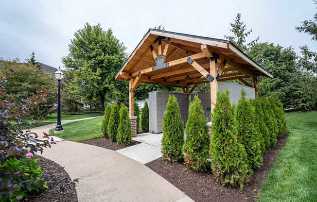 A wooden pavilion is surrounded by green shrubs and a concrete walkway.