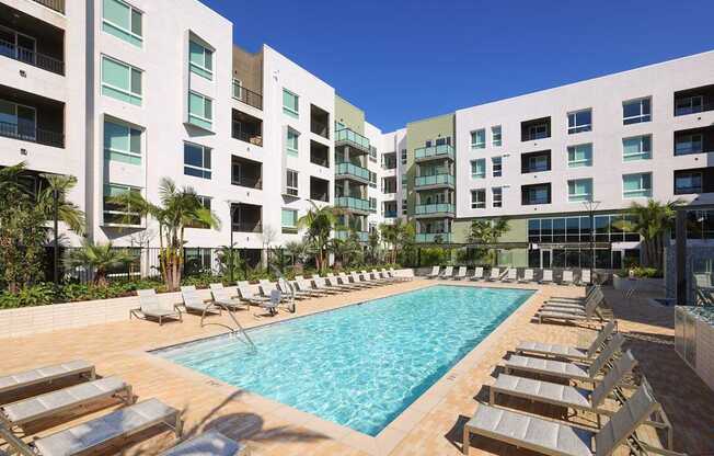 A swimming pool surrounded by sun loungers in front of apartment buildings.