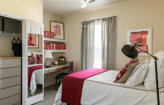 a bedroom with a bed with a red blanket and pillows and a desk with a computer