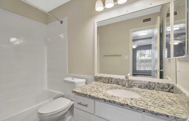 A well-lit bathroom featuring a white bathtub with a shower, a modern sink with a granite countertop, and a large mirror. Natural light streams in from a window in the background, highlighting the contemporary design and neutral color palette. The space is clean and organized, creating a welcoming atmosphere.