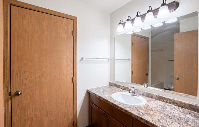 A bathroom with a sink, mirror, and wooden door. Fargo, ND Park Place Apartments