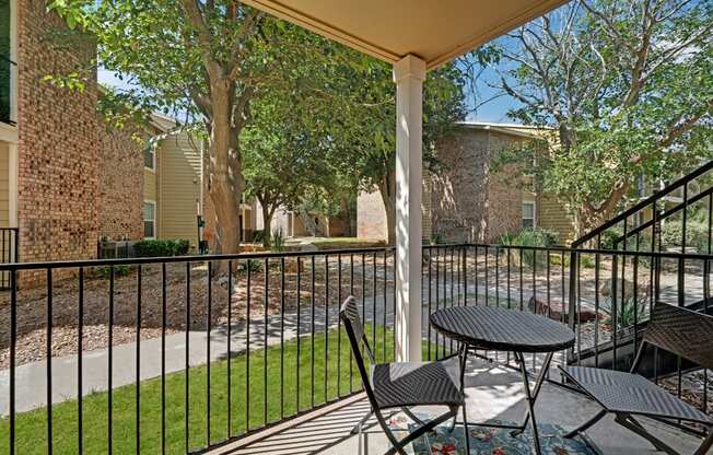A private patio here at The Bradford with a black metal railing, a small outdoor table with seating, and a covered area for shade. A peaceful courtyard view with mature trees, green landscaping, and walkways creates a calm and relaxing outdoor setting.