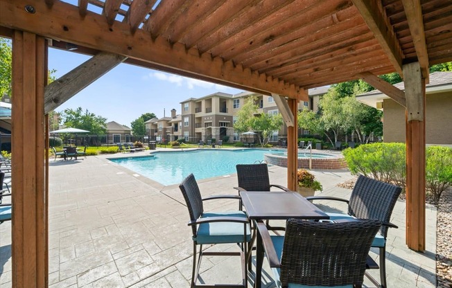 A wooden pergola over a table with chairs and a pool in the background.