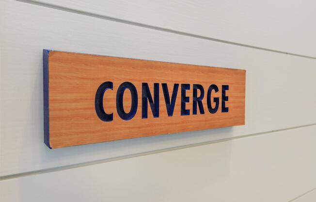 A wooden sign with the word "CONVERGE" displayed in bold, black letters against a light-colored wooden backdrop, mounted on a softly textured wall. The design emphasizes simplicity and clarity.