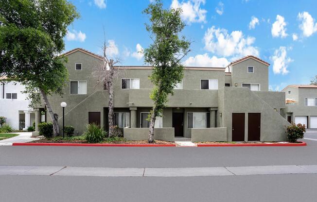 A modern, two-story apartment building with green exterior walls. The building features multiple windows and entrance doors, surrounded by small shrubs and trees. The sky is bright with fluffy clouds, and there is a paved path leading to the entrance, creating a welcoming atmosphere.