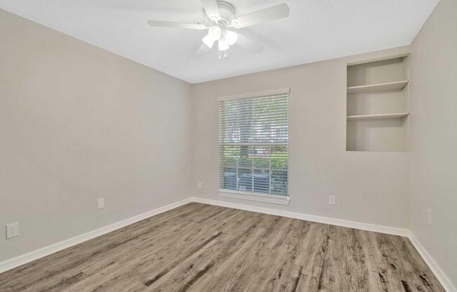 Empty room with light-colored walls, a ceiling fan, and a window with closed blinds. There is a built-in shelf on the right wall. The floor is composed of light wood laminate, creating a bright and spacious atmosphere.