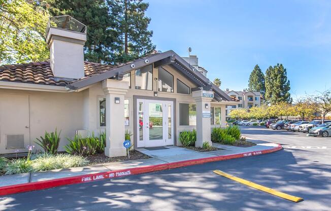 A modern building entrance with large glass doors, surrounded by greenery. The structure features a tiled roof and is set in a parking area, with several parked cars visible in the background. Bright blue skies and trees add to the welcoming atmosphere of the scene.
