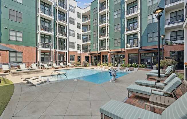 A large swimming pool surrounded by lounge chairs and umbrellas in front of apartment buildings.