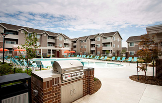 A bbq grill is in the foreground of a swimming pool surrounded by apartment buildings.