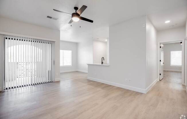 A spacious room with a ceiling fan and light fixture, hardwood flooring, and a window with blinds.