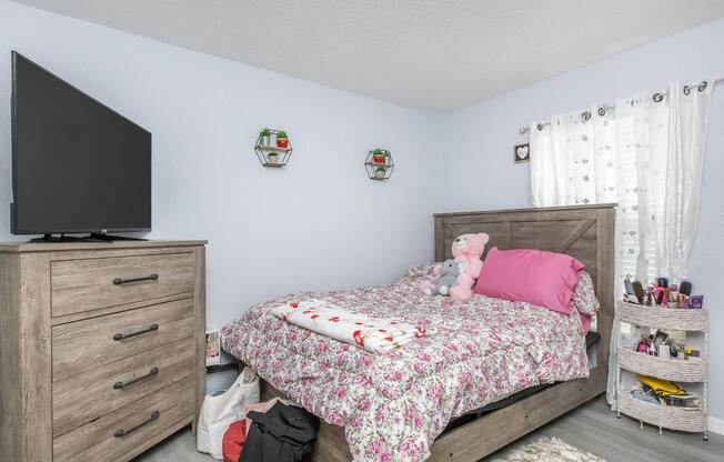 A cozy bedroom featuring a wooden bed with floral bedding, a pink pillow, and stuffed animals. There's a dresser with a television on top, alongside wall-mounted shelves with decorative items. A window with sheer curtains lets in light, and a small makeup organizer sits beside the bed.