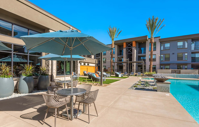 A patio with a table and chairs is surrounded by a pool and buildings.