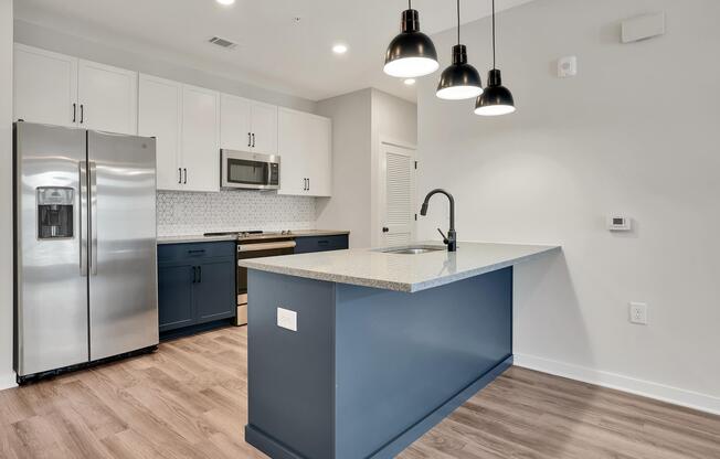 A modern kitchen featuring two-tone cabinetry with white upper cabinets and dark blue lower cabinets. It includes a stainless steel refrigerator, microwave, and a granite countertop with a sink. Three black pendant lights hang above the countertop, and there is a light wood flooring throughout.