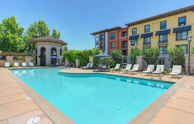 A swimming pool surrounded by lounge chairs and umbrellas in front of a multi-story apartment building.