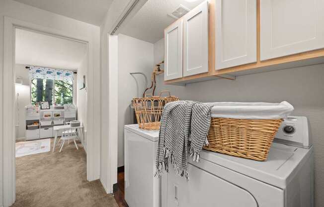 A laundry room with a washer and dryer, a basket on top of the dryer, and a window with a view of trees at Clackamas Trails Apartments, Oregon, 97222