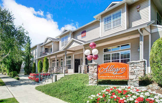 A welcoming leasing office exterior here at Allegro featuring a stone monument sign with the Allegro Apartment Homes name, manicured flower beds, and a sidewalk leading to a two-story building with neutral siding, large windows, covered entryways, and landscaped greenery under a clear blue sky.