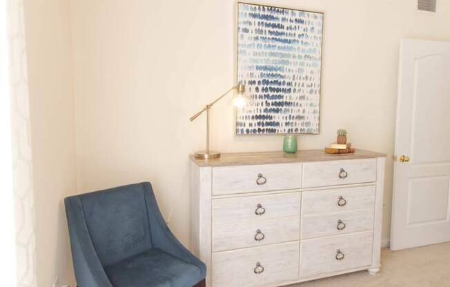 A blue chair sits in front of a white dresser with a framed artwork on the wall.