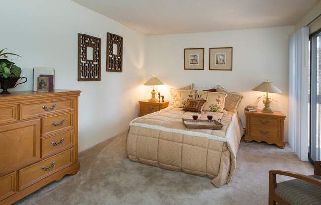 A comfortable bedroom here at Eagle Ridge featuring a neatly made bed with layered neutral bedding, matching wood nightstands with soft table lamps, framed wall art above the headboard, plush carpeted flooring, a spacious dresser for storage, and a sliding glass door that brings in natural light and connects the room to an outdoor area.