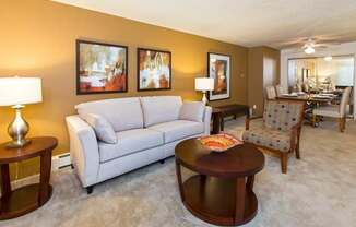 A bright living room here at Eagle Ridge featuring a light gray sofa paired with a round wooden coffee table, matching side tables with modern lamps, and abstract wall art along a warm neutral accent wall, with plush carpeting and an open layout that flows into a dining area furnished with upholstered chairs and a rectangular table.