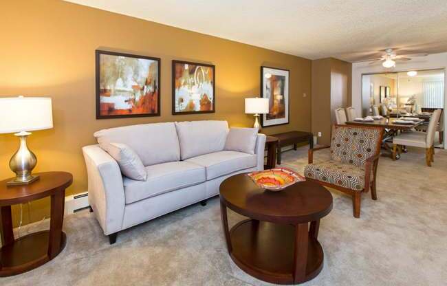 A bright living room here at Eagle Ridge featuring a light gray sofa paired with a round wooden coffee table, matching side tables with modern lamps, and abstract wall art along a warm neutral accent wall, with plush carpeting and an open layout that flows into a dining area furnished with upholstered chairs and a rectangular table.
