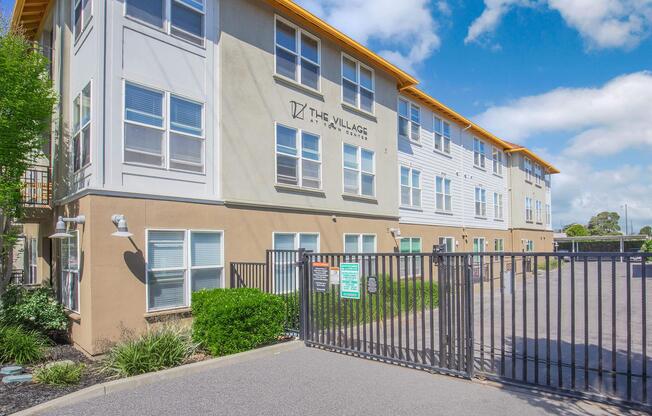 Exterior view of a multi-story residential building named "The Village," featuring large windows and a security gate at the entrance. The surrounding area includes landscaped greenery, with a clear sky in the background.
