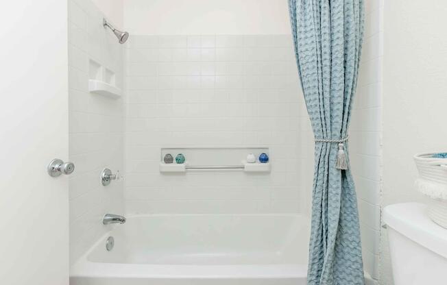 A clean and modern bathroom featuring a white tiled shower area with a bathtub. A light blue shower curtain hangs on the rod, and there are grab bars for assistance. White shelves on the wall hold toiletries. A white toilet is positioned near the shower area, completing the minimalist aesthetic.