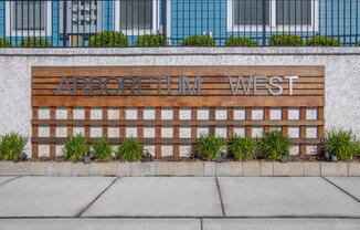 A low wooden sign displaying "ARBORETUM WEST" in metallic letters, set against a modern building facade. The sign is surrounded by low green shrubs and placed on a concrete pathway, creating a contemporary and inviting entrance.