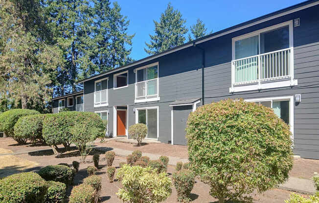 A grey building with a red door and windows surrounded by green bushes.