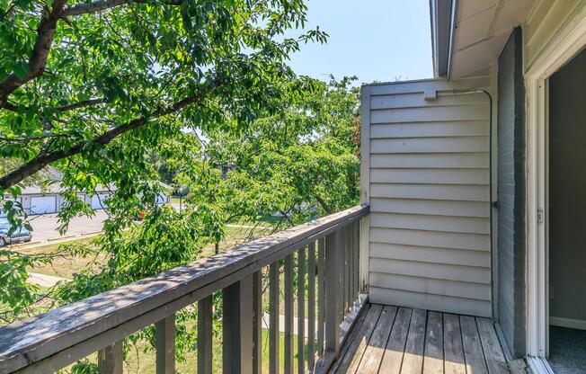 View from a balcony looking out onto greenery and a pathway. The wooden railing frames the image, with leaves from nearby trees creating a natural setting. A corner of a building is visible, suggesting a residential area. The scene is bright and inviting, with clear blue skies above.