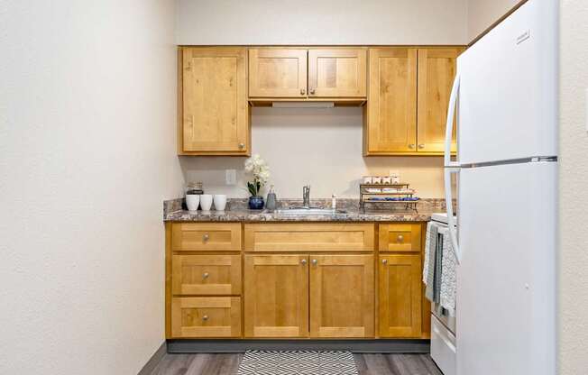 A kitchen with wooden cabinets and a white refrigerator A row of apartment buildings with green lawns in front A bedroom with a bed, nightstand, and window at Wildflower, Kennewick, WA 99336..