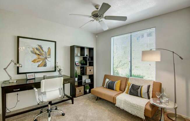A versatile home office and guest space here at Mirador featuring soft neutral walls, plush carpeting, and a ceiling fan overhead. A sleek dark wood desk with a modern chair sits beneath framed artwork, while a cozy sofa with accent pillows and a throw rests beside a floor lamp and large window with blinds that invite natural light into the room.