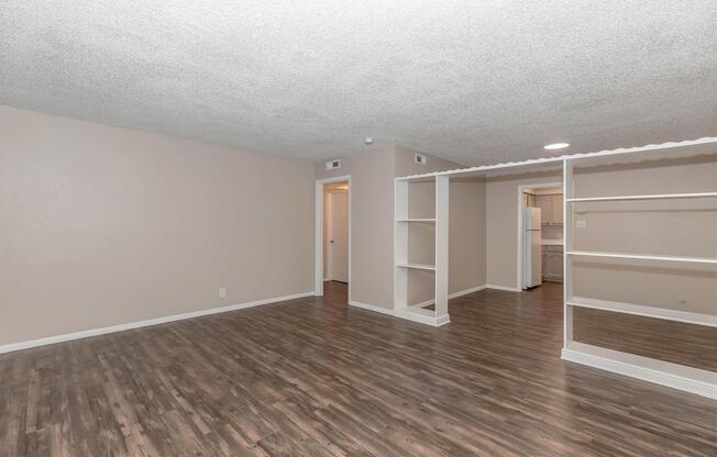 Spacious living area with a textured ceiling and laminate wood flooring. Light-colored walls and an open layout leading to a kitchen area with a refrigerator visible in the background. A narrow shelf divides the space, enhancing the room's airy feel. Doorways lead to additional rooms.