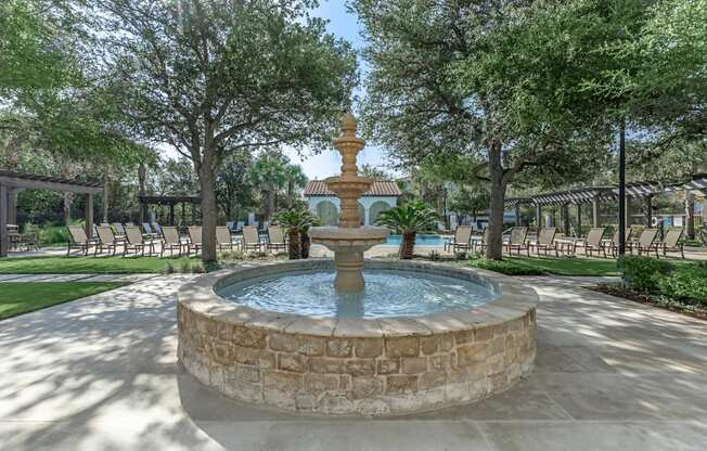 A fountain in the middle of a park with trees and chairs around it.