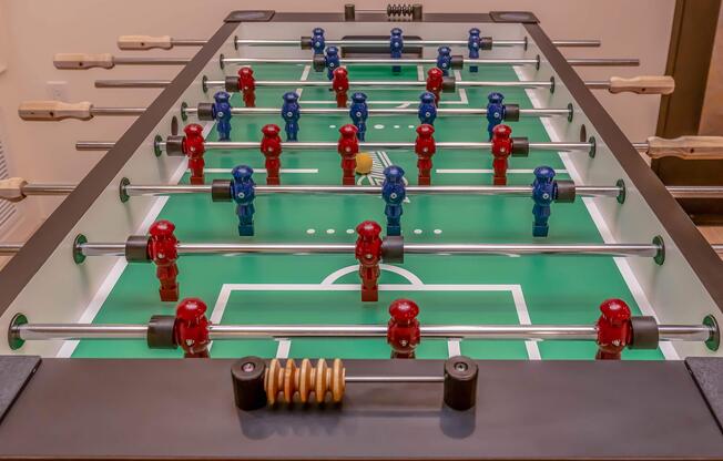 A foosball table featuring a green playing surface with miniature players in red and blue formations. The table has metallic rods, and a yellow ball is positioned at the center, ready for play.