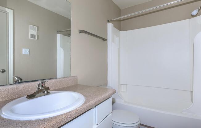 A clean and simple bathroom featuring a white bathtub and shower combo, a sink with a faucet on a beige countertop, and a toilet. The walls are painted in a soft beige color, and there is a large mirror above the sink reflecting the space. A towel bar is mounted on the wall.