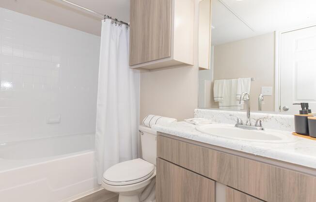 A clean, modern bathroom featuring a white bathtub with an attached shower curtain, a sleek vanity with a sink, and wooden cabinetry. Fresh towels are neatly arranged, and there's a mirror above the sink. The space is well-lit and has a neutral color palette, creating a bright and inviting atmosphere.