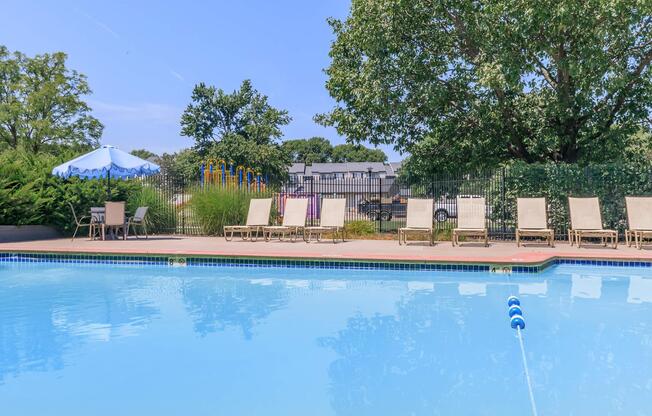 A clear swimming pool surrounded by lounge chairs, with a blue and white umbrella in the background. The pool's surface reflects the sky and trees, while a fenced area with playground equipment is visible in the distance.