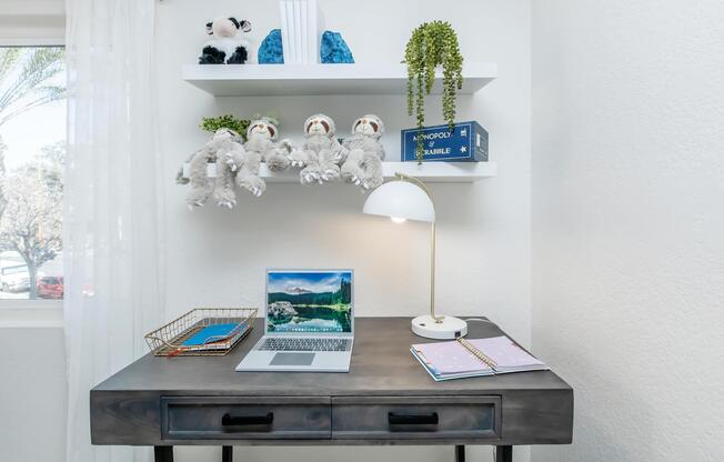 A modern workspace featuring a wooden desk with a laptop displaying a scenic landscape, a decorative basket, and a notebook. Above the desk, there are shelves containing plush toys, decorative items, and a hanging plant, with natural light filtering in through a nearby window.