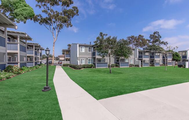A peaceful residential area featuring two-story apartment buildings set amidst well-maintained lawns and trees. A paved walkway leads through the green space, with light posts adding to the inviting atmosphere under a clear blue sky.