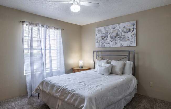 A bedroom with a bed, pillows, a ceiling fan, and a painting on the wall.