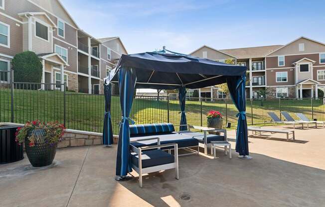 A gazebo with a blue canopy is situated in a courtyard with lounge chairs and potted plants.