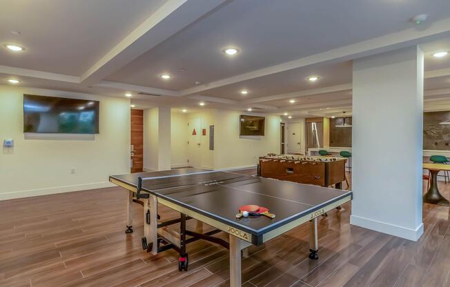 A spacious game room featuring a ping pong table and a foosball table. The room has modern decor with wooden flooring, recessed lighting, and large flat-screen TVs mounted on the walls. There are also green chairs and a sleek table in the background, creating a vibrant recreational space.