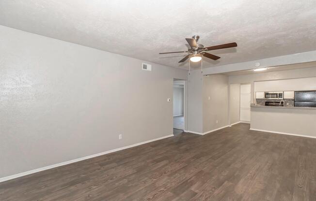 A spacious, empty living room featuring light gray walls, a ceiling fan, and hardwood-like flooring. An open layout connects to a kitchen area in the background, which includes modern appliances and a bar counter. Natural light brightens the space, creating a welcoming atmosphere.