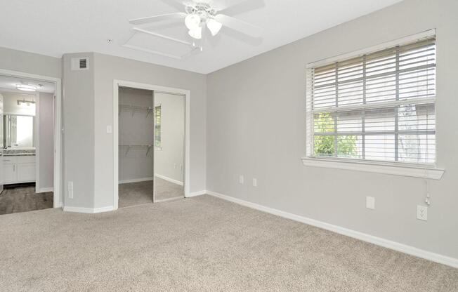 A spacious, neutral-colored room with carpeted flooring. It features a ceiling fan, a window with blinds, and a closet with an open door. There is an adjacent doorway leading to a bathroom area with a countertop visible. The natural light creates a bright and inviting atmosphere.
