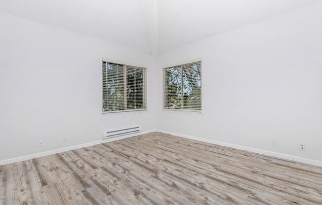A bright, empty room with white walls and two large windows featuring blinds. The flooring is light wood, and the ceiling has a vaulted design. Natural light floods the space, casting soft shadows on the floor.