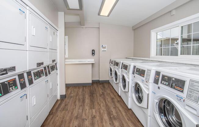 A modern laundry room featuring several stacked washing machines and dryers lined up against the wall. The space includes a folding area with a countertop, is well-lit with overhead lighting, and has neutral-colored walls. Large windows allow natural light to enter the room.