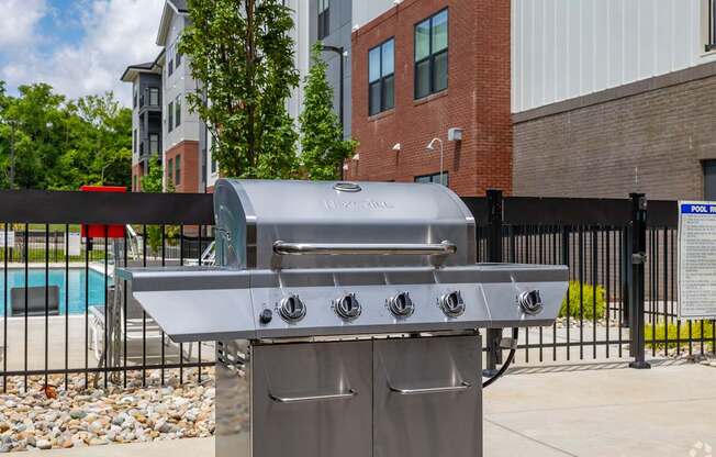 A silver gas grill with four burners is in front of a black fence.