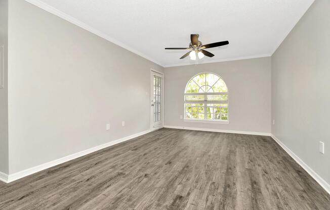 A spacious, empty living room featuring light gray walls, modern wood-like flooring, and a ceiling fan. Natural light streams in through a large arched window with white trim, and there is a door leading to an outdoor space. The overall ambiance is bright and inviting.